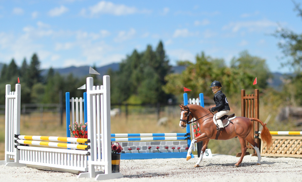 A chestnut model pony seen in a jumper course with a doll rider and multiple obstacles.