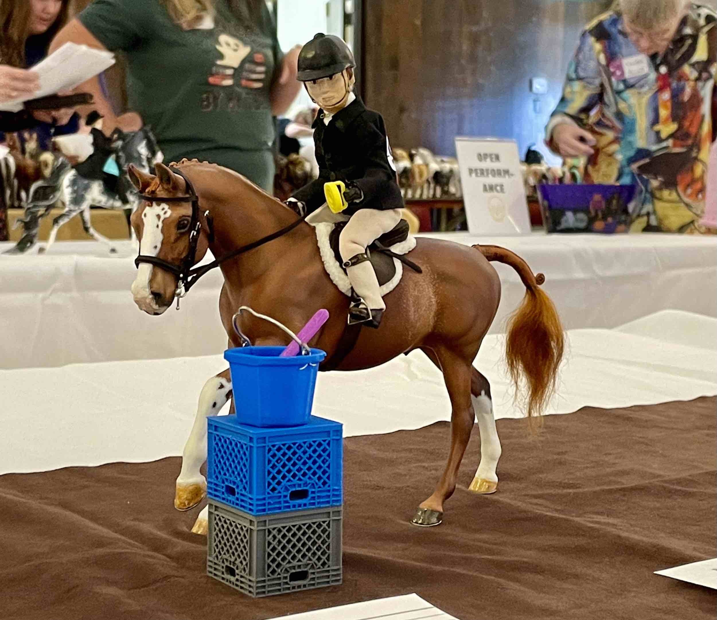 A chestnut model pony with a doll rider is dropping a currycomb in a bucket as part of a speed game